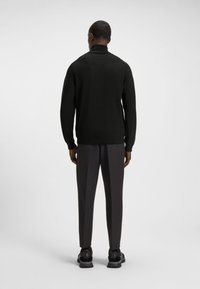 Juodas megztinis iš ribos mezgimo su aukštu apykakliu, dėvimas kartu su tamsiai pilkomis kelnėmis. Išvaizdą papildo juodi batai su tekstūruotomis pado dalimis.