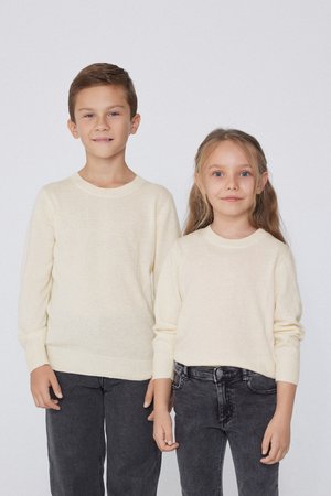 Zwei Kinder stehen nebeneinander, beide tragen cremefarbene Pullover und dunkle Jeans, vor einem schlichten, hellen Hintergrund.