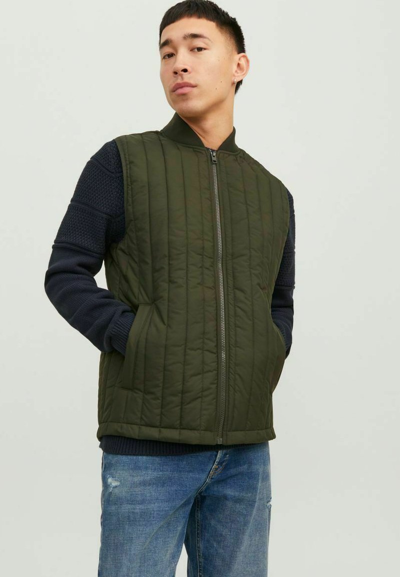 Junger Mann mit kurzen Haaren, der eine grüne gesteppte Weste über einem marineblauen Pullover und blauen Jeans trägt, steht mit den Händen in den Westentaschen.