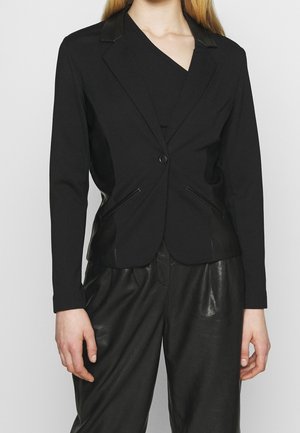 Frau trägt einen figurbetonten schwarzen Blazer mit Reißverschlusstaschen über einem schwarzen Top, kombiniert mit locker sitzenden schwarzen Lederhosen vor einem schlichten Hintergrund.