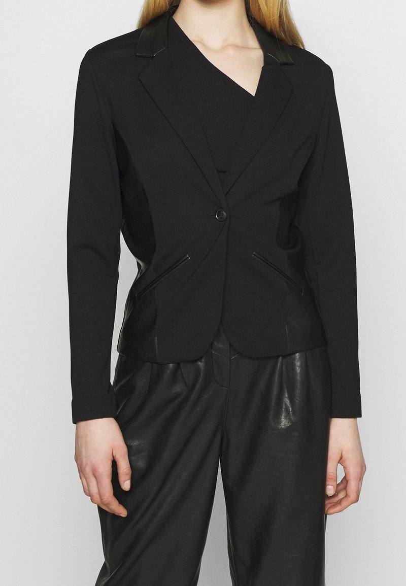 Femme portant un blazer noir cintré avec des poches zippées sur un top noir, associé à un pantalon en cuir noir ample sur un fond uni.