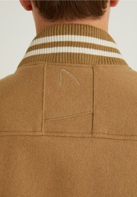 Personne portant un manteau marron chameau avec un col côtelé aux rayures blanches et marron, photographiée de dos montrant les détails des coutures du manteau.