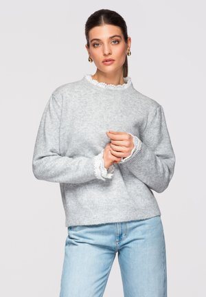 Ung kvinde med opsat hår, iført gyldne øreringe i hoop- stil, lysegrå sweater med blondekant og lyseblå jeans, stående mod en hvid baggrund.