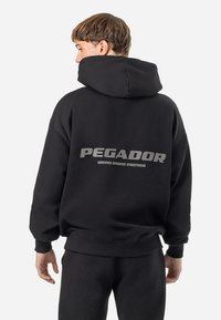 Mustat hupparit, jossa on löysä istuvuus, takana suuri harmaa "PEGADOR" -logo. Valmistettu pehmeästä materiaalista, jossa on ribattut hihansuut.