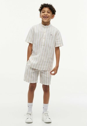 Ragazzo giovane sorridente, indossa una maglietta a maniche corte beige e bianca a righe e pantaloncini coordinati, calzini bianchi e scarpe da ginnastica bianche, in piedi in modo casuale.