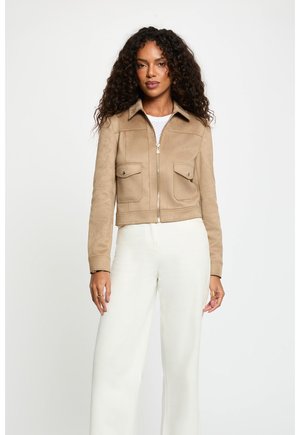 Mujer con cabello rizado que lleva una chaqueta marrón con cremallera y bolsillos con solapa, y pantalones blancos de pierna ancha, de pie frente a un fondo liso.