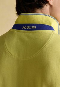Žlté polo tričko s textúrovanou tkaninou, s námorníckym modrým golierom a značkou "JOULES" vyšitou bielou farbou na chrbte.