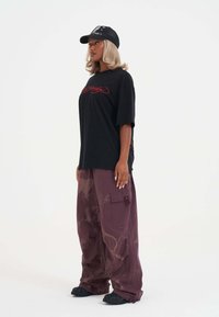 Sort oversized t-shirt med rød grafik, parret med løse lilla bukser med et subtilt mønster og sidelommer. Sorte sneakers og kasket.