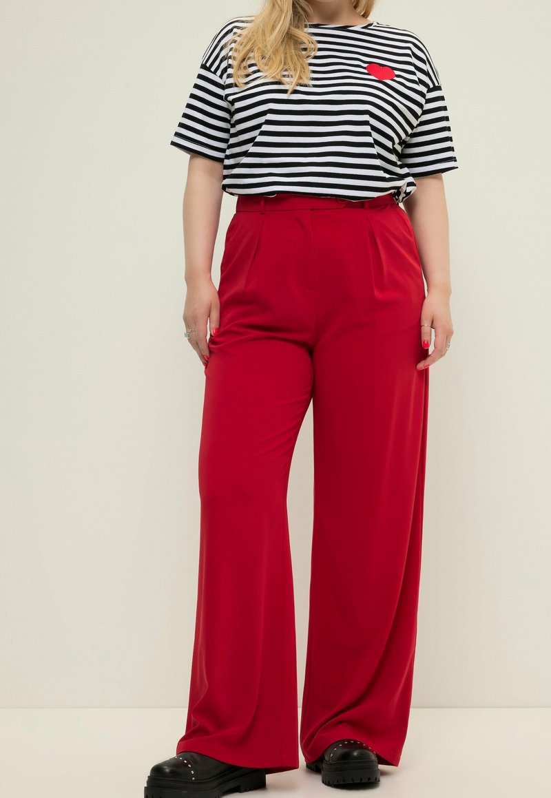 Czerwone spodnie z szerokimi nogawkami i wysokim stanem, w zestawieniu z czarno-białą koszulką w paski z małym czerwonym serduszkiem oraz masywnymi czarnymi butami.