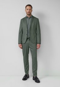 Grüner strukturierter Anzug mit einem taillierten Blazer, maßgeschneiderten Hosen und einem passenden Hemd, das eine sanfte Oberfläche aufweist, sowie schwarzen Loafers.