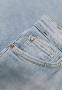 Světle modré džíny z denimu s malou přední kapsou, zlatým prošitím a měděnými knoflíkovými detaily. Tkanina má hladkou texturu.