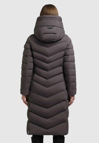 Donna che indossa un lungo cappotto imbottito grigio scuro con grande cappuccio, vista di spalle su uno sfondo chiaro e semplice.