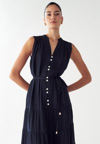Námornícky bezrukávový šaty s výstrihom do V, predným zapínaním na gombíky, zúženým pásom a vrstvenou sukňou; má jemnú textúru a zlaté akcenty na stužke.