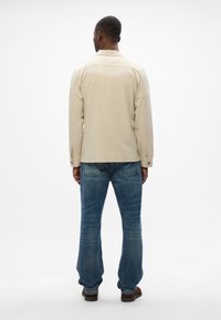 Μπεζ πουκάμισο με γιακά συνδυασμένο με μπλε τζιν. Το πουκάμισο έχει λεία υφή, ενώ τα τζιν παρουσιάζουν μουντζουρωμένο σχέδιο και ίσια γραμμή.