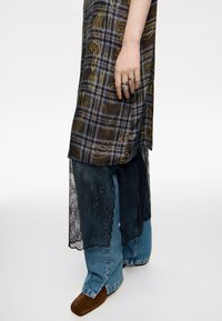 Largo vestido de cuadros con patrones amarillos y azules, superpuesto sobre encaje oscuro y mezclilla, con un borde de encaje en forma de ondas y zapatos marrones.