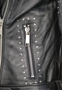 Sortlæderjakke med en sølvfarvet lynlås og nitter. Overfladen har en glat tekstur med metalnitter langs siden.