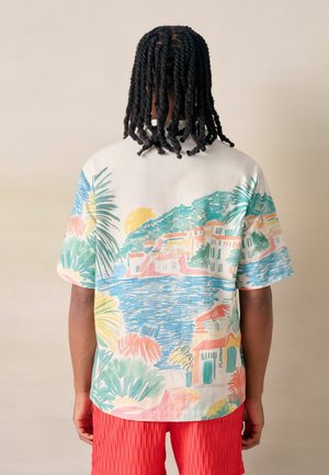 Mann mit gedrehten schwarzen Haaren, der ein Hemd mit pastellfarbenem tropischem Muster und strukturierte rote Shorts trägt, steht vor einem einfachen hellen Hintergrund.