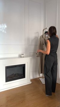 Un trench coat bej cu butoane duble, expus pe un suport de haine într-o cameră luminoasă, cu un șemineu alb și podea din lemn.