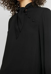 Blusa nera con colletto legato, maniche lunghe e bottoni decorativi sulle spalle. Tessuto liscio con finitura opaca.