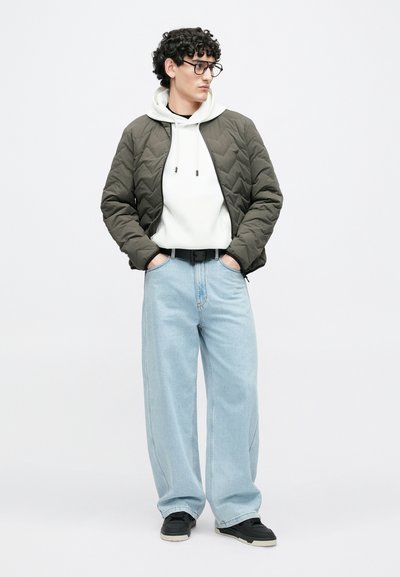 Prešívaná olivovo zelená bunda, biela mikina s kapucňou a šnúrkami, svetlomodré voľné džínsy, čierne tenisky s bielymi podrážkami a čierne okuliare.