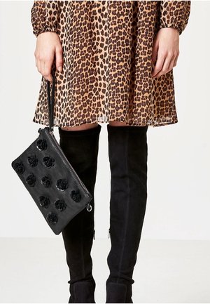 Mani che tengono una pochette nera con applicazioni floreali, abito a stampa leopardata, e stivali neri in camoscio sopra il ginocchio visibili dalla metà coscia in giù.