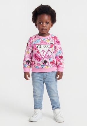 Bambino piccolo con felpa rosa colorata con logo "Guess", jeans azzurri chiari e sneakers bianche, in piedi contro uno sfondo semplice.