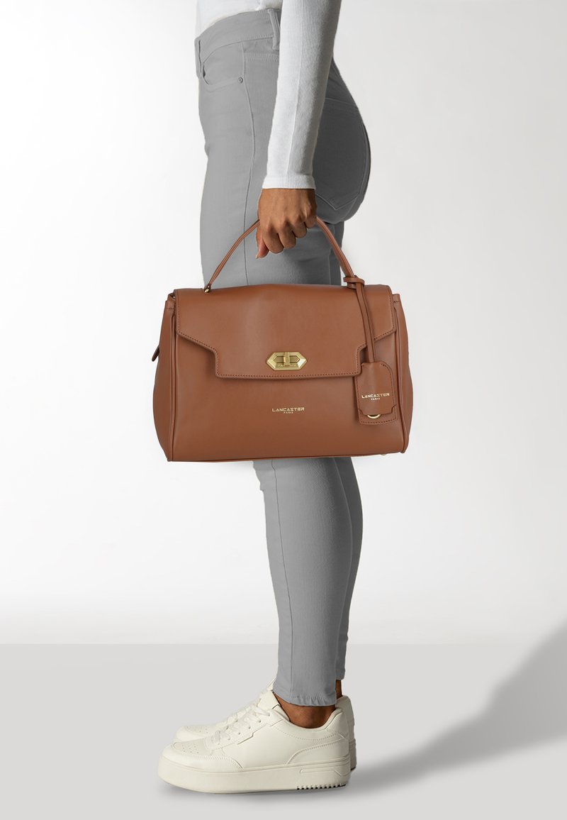 Borsa a mano in pelle marrone con una forma strutturata, hardware dorato, chiusura a patta e un'etichetta attaccata. La borsa ha una texture liscia e minimi dettagli.