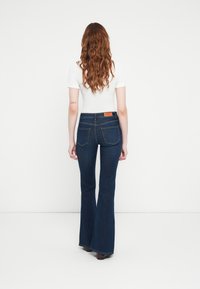 Jean évasé en denim bleu foncé avec une taille haute, comportant deux poches arrière et une étiquette marron sur la ceinture. Haut ajusté blanc.