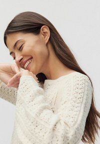 Sweter robiony na drutach w kolorze kremowym, z delikatnym wzorem ażurowym, okrągłym dekoltem i długimi rękawami z ściągaczami. Miękka tekstura na tle.