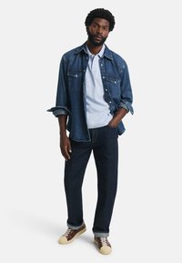 Jeansowa kurtka na jasnoniebieskim polo, w zestawie z ciemnymi dżinsami i brązowymi trampkami. Posiada detale z guzikami i podwinięte rękawy.