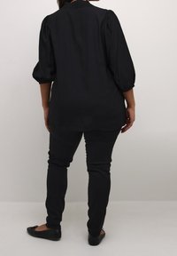 Blouse noire à manches bouffantes, col texturé et coupe ample, associée à un pantalon noir ajusté et des chaussures plates. Vue de dos.