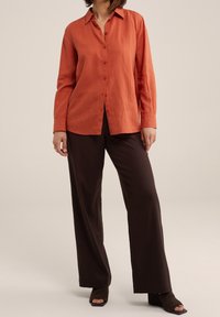 Ein leinen Hemd in rostiger Farbe mit langen Ärmeln und klassischem Kragen. Kombiniert mit dunkelbraunen, weiten Hosen und Absatzschuhen.