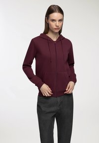 Felpa a zip color bordeaux con tasca frontale, cordoncini e polsini a costine, abbinata a pantaloni grigi a vita alta. Tessuto dalla texture liscia.