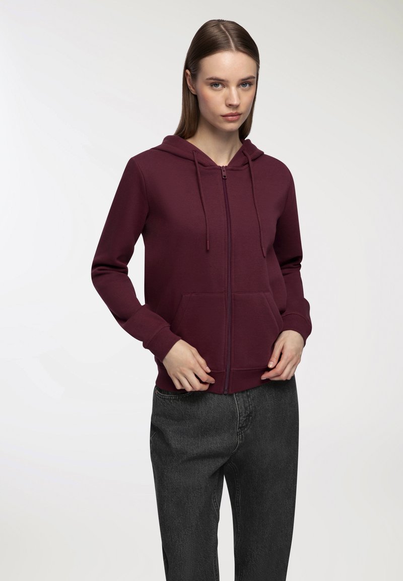 Felpa a zip color bordeaux con tasca frontale, cordoncini e polsini a costine, abbinata a pantaloni grigi a vita alta. Tessuto dalla texture liscia.