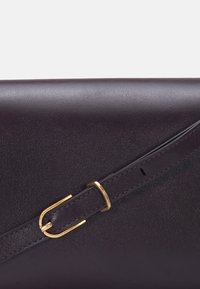 Sac en cuir violet foncé avec une fine bandoulière réglable dotée d'une petite boucle rectangulaire en or, vue rapprochée de la texture et des coutures.