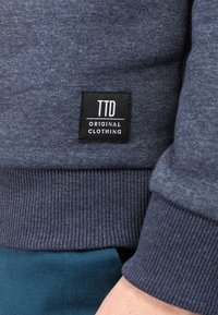 Felpa blu navy con polsini e orlo a costine, con un'etichetta nera con il testo "TTD ORIGINAL CLOTHING" sul lato. Tessuto liscio.
