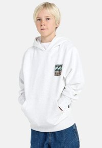 Blond jongetje met sproeten, gekleed in een witte hoodie met een klein groen en beige logo op de borst en blauwe jeans, met de handen in zijn zakken.