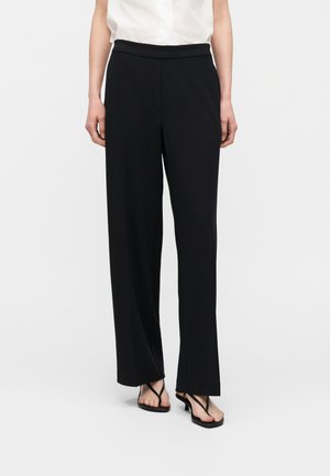 Persona che indossa pantaloni neri a gamba larga, top bianco senza maniche e sandali neri con tacco, in piedi su uno sfondo bianco.