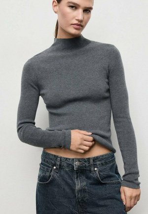 Donna che indossa un maglione grigio a collo alto attillato e jeans neri sbiaditi, mentre tiene l'orlo del maglione con una mano.
