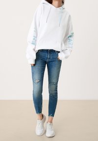 Sweat à capuche blanc avec coupe oversize, manches longues et accents de texte pastel. Associé à un jean skinny bleu déchiré et des baskets blanches.