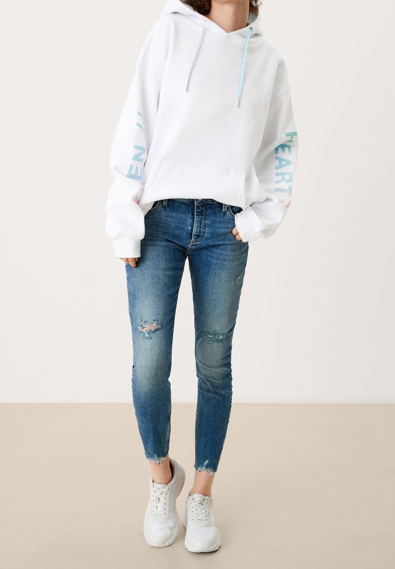 Sweat à capuche blanc avec coupe oversize, manches longues et accents de texte pastel. Associé à un jean skinny bleu déchiré et des baskets blanches.