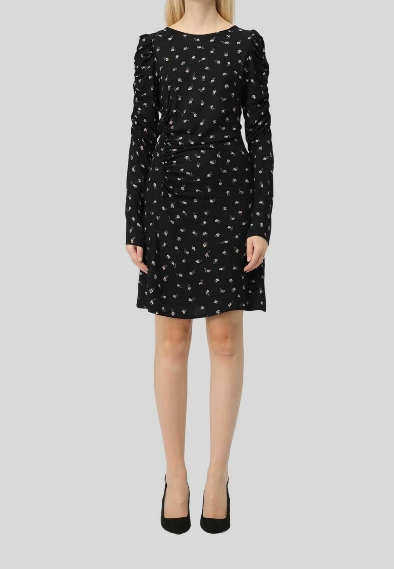 Zwart lang mouwjurk met bloemenpatroon, gerimpelde zijkanten en ronde halslijn. De stof is glad; gecombineerd met zwarte hoge hakken.