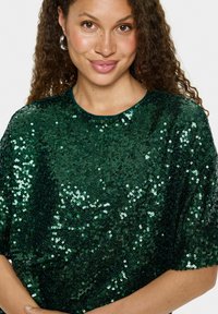Top en sequins verts avec une coupe décontractée, un col rond et des manches courtes. Texture lisse avec des sequins réfléchissants créant un effet scintillant.