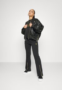 Čierna bomber bunda so lesklým povrchom a zlatými zipsami, skombinovaná s čiernymi nohavicami so bočnými zipsami a malým zlatým emblémom. Biele tenisky.