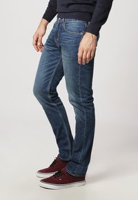 Blue denim jeans with a slim fit, featuring a mid-rise waist, five pockets, and contrasting stitching. The model wears burgundy sneakers.