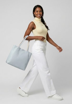 Usmívající se žena kráčející, oblečená v bezrukávovém béžovém roláku, bílých kalhotách a bílých teniskách, nese velkou světle modrou tašku přes rameno.