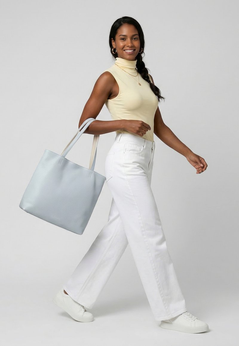 Femme souriante marchant, portant un col roulé sans manches beige, un pantalon blanc, des baskets blanches, et portant un grand sac cabas bleu clair à l'épaule.