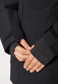 Black outdoor jacket with a smooth texture, featuring Velcro cuffs and a close-fitting design. Hands are clasped together at the waist.