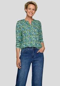 Mehrfarbige Bluse mit langen Ärmeln und V-Ausschnitt, die grüne und blaue geometrische Muster aufweist. Kombiniert mit hochgeschnittenen, dunklen Jeans.