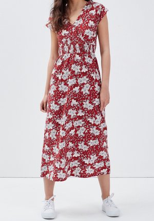 Femme portant une robe midi rouge à imprimé floral blanc, manches courtes, taille smockée, associée à des baskets blanches sur un fond uni.